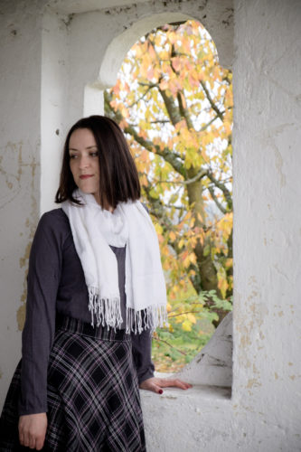Rock, Shirt und Schal ohne Fellweste. Lehnt am Fensterbrett, draußen sieht man das Herbstlaub eines Baumes