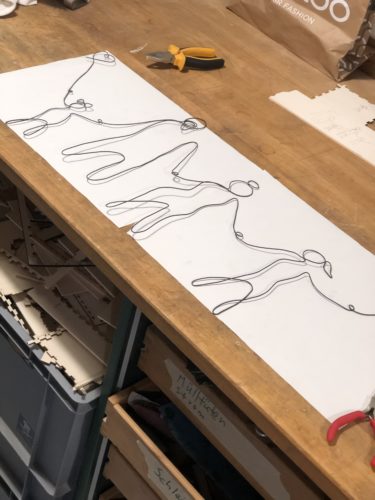 Der Draht ist fertig zum Familienportrait gebogen und liegt auf einem weißen Papier auf der Werkbank