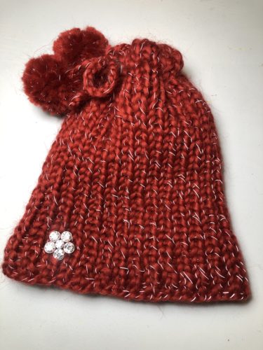 Der Loop ist oben mit der Kordel zusammengezogen worden, sodass eine rote Mütze entstanden ist. Die Kordelenden sind zu einer Schleife gebunden, sodass die beiden Bommel oben an der Mütze sind. 