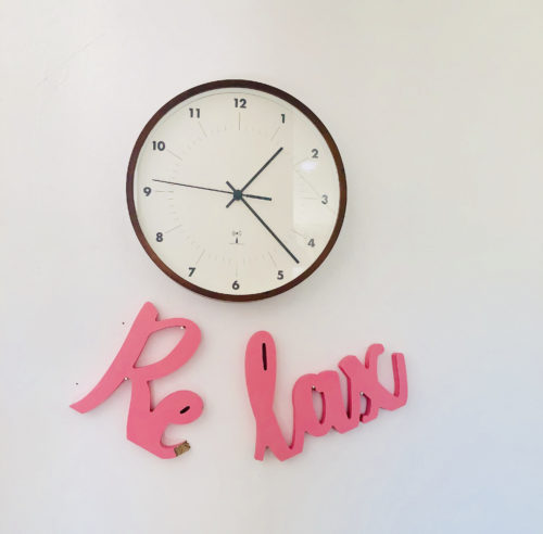 Uhr an der Wand. Darunter ein zerbrochener Schriftzug aus Holz "Relax"
