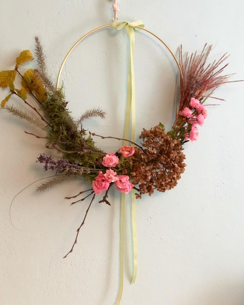 Der fertige Kranz hängt an der Wand. die untere Hälfte des goldenen Rings ist mit Gräsern, Moos, Zweigen und Blüten umwickelt. Diesmal aus der Nähe fotografiert. Die Rosen in rosa und das Gras mit leichtem Rotschimmer ergänzen sich farblich gut.