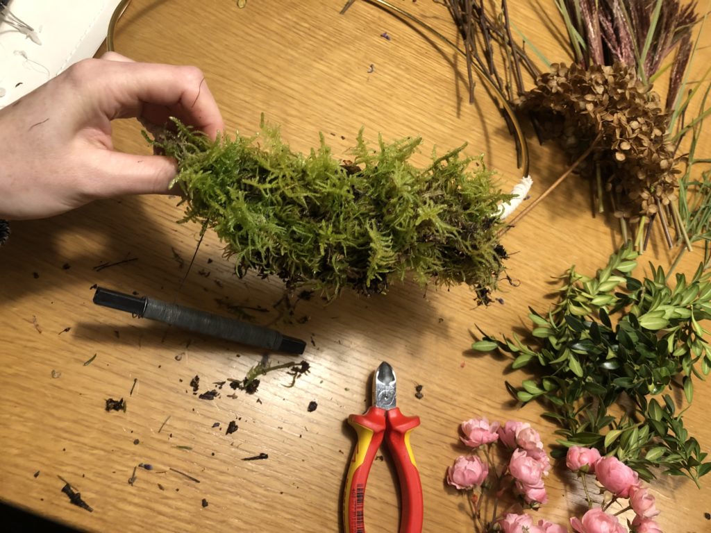 Der Kranz ist zu 1/3 mit Moos umwickelt. Weiteres Zubehör, wie Buchs und kleine Äste, liegt auf dem Tisch daneben. 