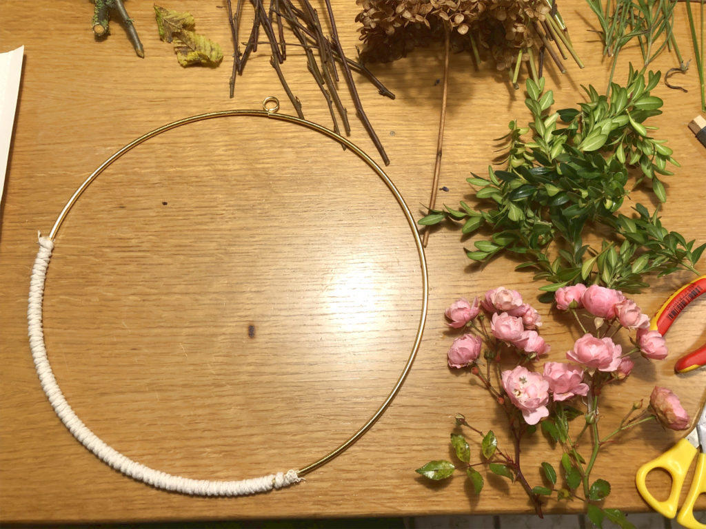 Der goldene Ring ist zu 1/3 mit naturfarbenem Seil umwickelt. Neben dem Ring auf dem Tisch liegen rosa Rosen, Zweige und Äste.
