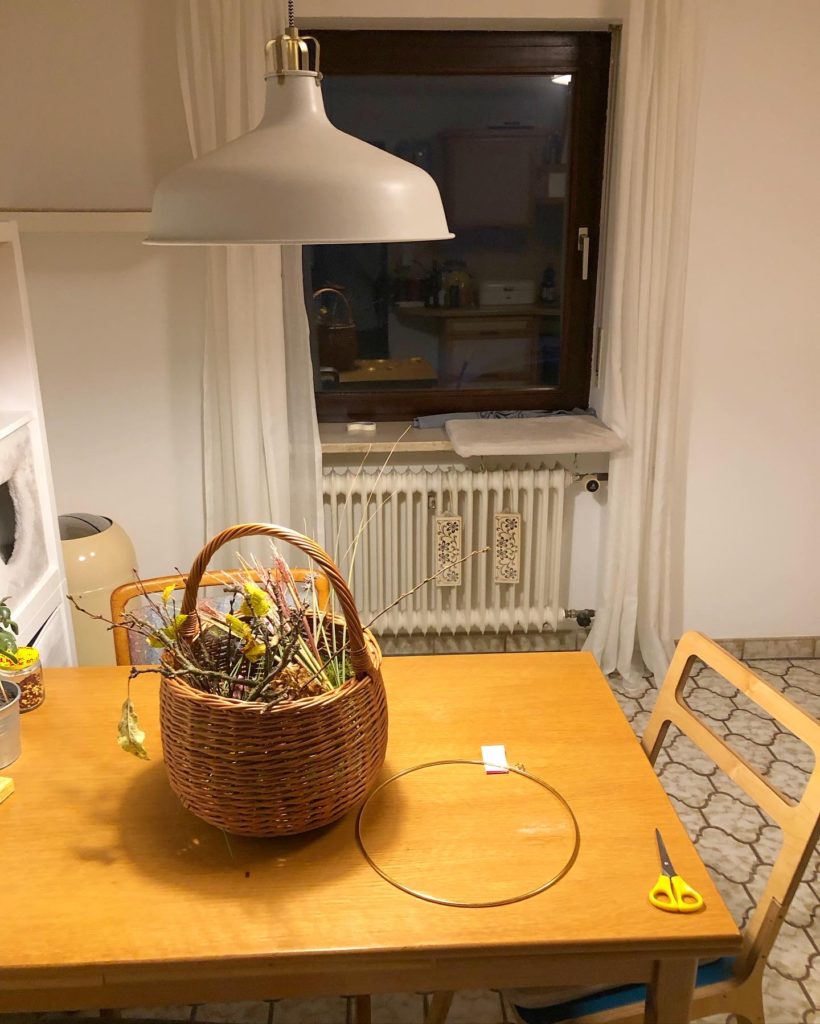 eine runder Korb steht auf dem Küchentisch gefüllt mit allerhand Gräsern, Ästen und Zweigen. Daneben liegen ein goldener Ring und eine gelbe Schere.