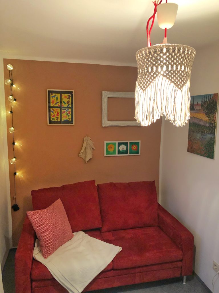 Makramee Lampenschirm aus naturfarbenem Seil hängt an der Decke eines kleinen Raumes. Das Licht im Lampenschirm ist an und das Makramee Muster kommt gut zur Geltung. Im Hintergrund steht vor einer altrosa Wand ein rotes Sofa. An der Wand hängen Bilder und links in der Ecke des Zimmers leuchtet eine Lichterkette. 