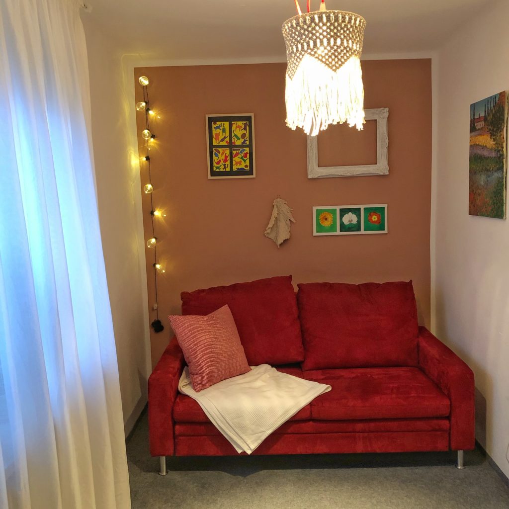 Makramee Lampenschirm aus naturfarbenem Seil hängt an der Decke eines kleinen Raumes. Das Licht im Lampenschirm ist an und das Makramee Muster kommt gut zur Geltung. Im Hintergrund steht vor einer altrosa Wand ein rotes Sofa. An der Wand hängen Bilder und links in der Ecke des Zimmers leuchtet eine Lichterkette. Links im Bild ist der weiße Vorhang vor dem Fenster zu sehen.