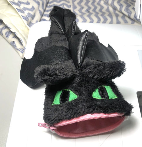 schwarze Tasche die mit Ohren, Flügeln, grünen Augen und Zacken die auf der Mitte entlang laufen den Drachen Ohnezahn stilisiert. Ein rosa Reißverschluss bildet die Taschenöffnung und gleichzeitig das Maul ohne Zähne. 