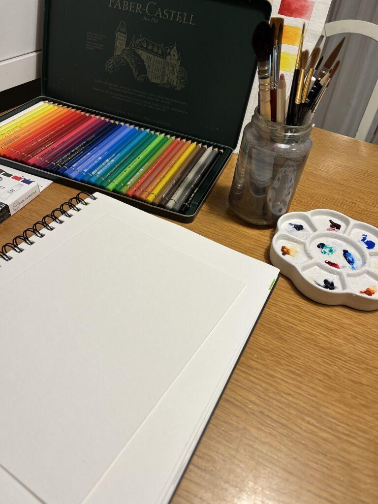 weißes Blatt Papiert und drum herum auf dem Tisch jede Menge Malzubehör, wie Farben, Pinsel und Buntstifte.